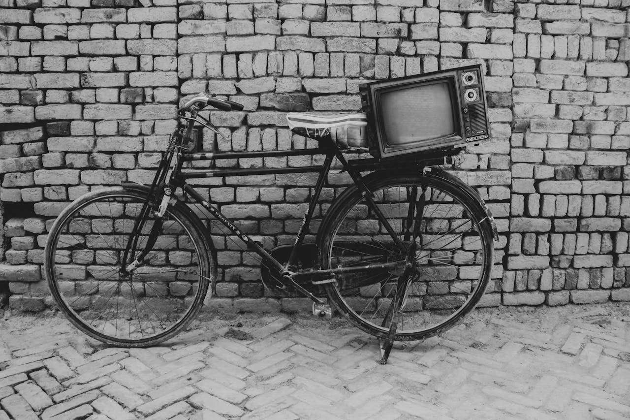 services-04 A vintage bicycle with an old television set mounted, set against a rustic brick wall in grayscale.