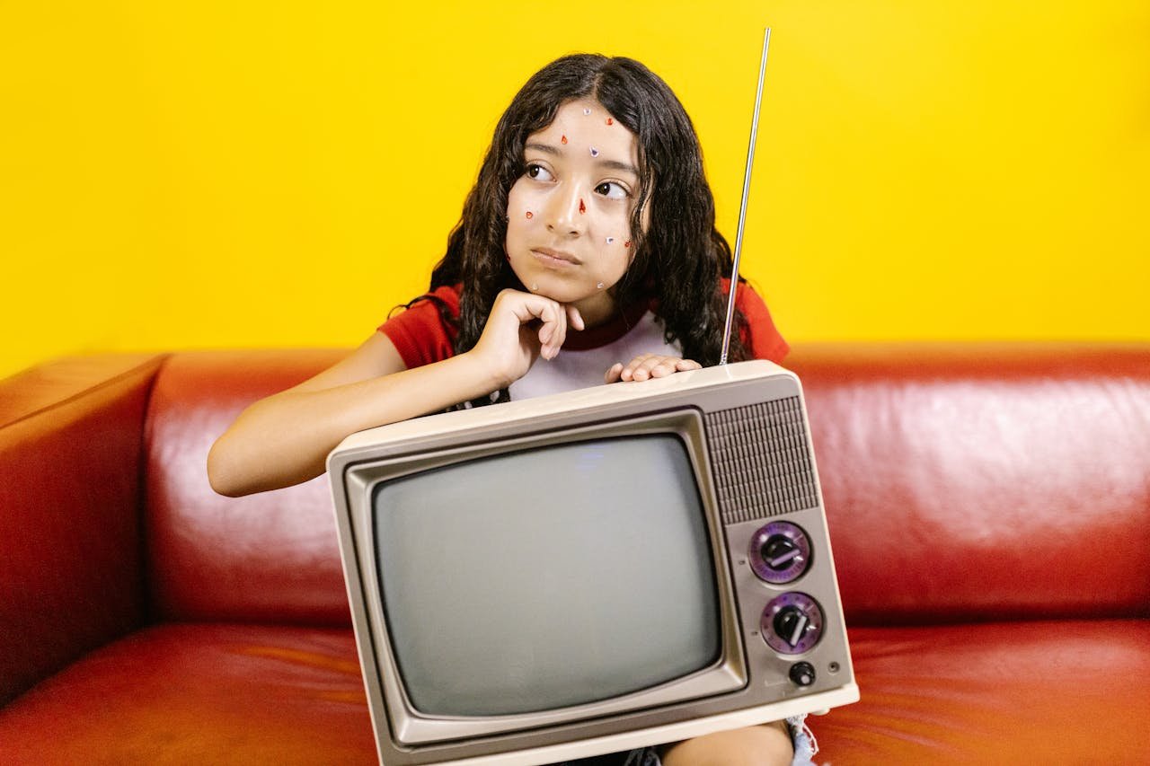 journey Woman with stickers on face posing with a vintage TV on a red couch with a bright yellow background.